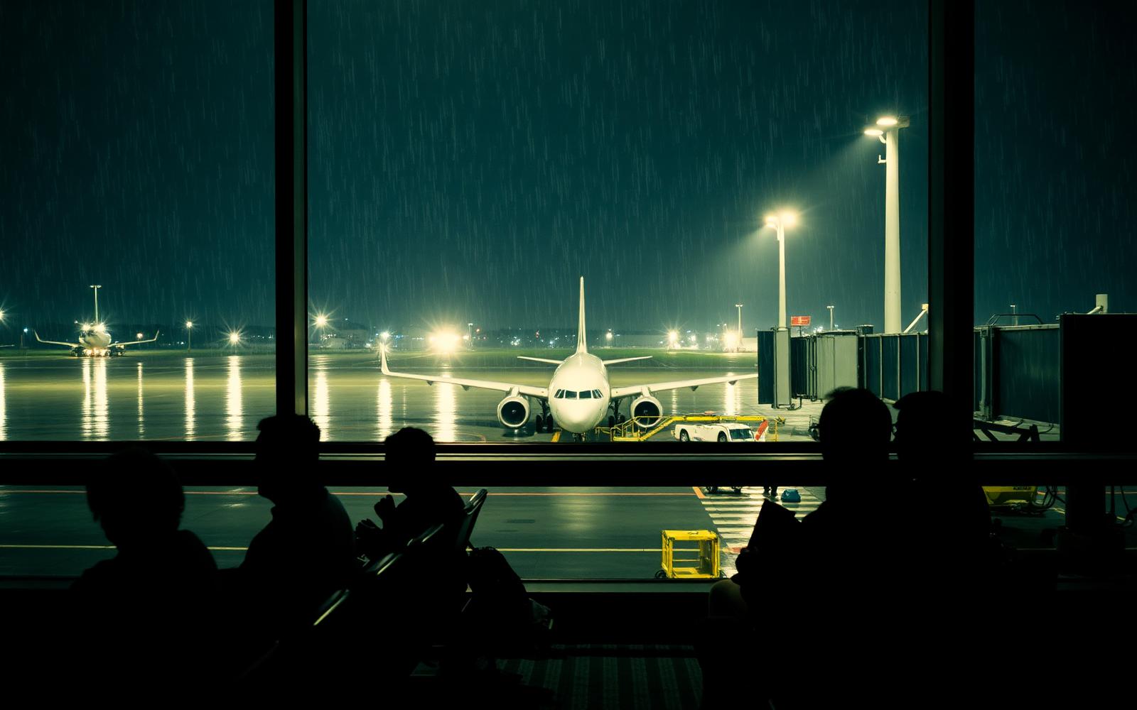 Passengers waiting at a gate at night as an aircraft stands on a rain-soaked apron